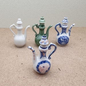 Könnte beinhalten: Vier kleine Keramik-Teekannen in verschiedenen Farben und Designs. Zwei sind weiß mit blauen und goldenen Akzenten, eine ist grün mit goldenen Akzenten und eine ist weiß mit rosa und blauen Akzenten.