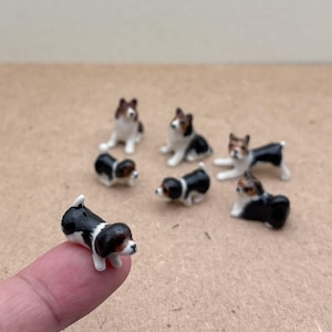 Op de afbeelding: Een set van acht miniatuurhondjes van keramiek. De beeldjes zijn geschilderd in zwart, wit en bruin. De honden staan in verschillende poses, waaronder zittend, staand en liggend.