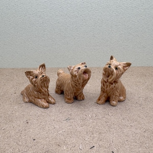 Puede incluir: Tres peque&ntilde;as figuritas de cer&aacute;mica de perros Yorkshire Terrier. Los perros son marrones y blancos y est&aacute;n en diferentes poses.