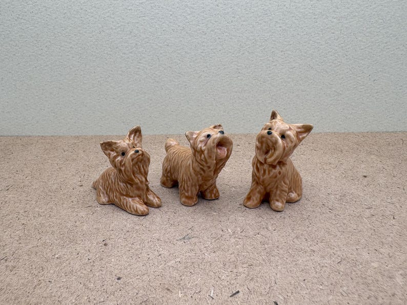 Puede incluir: Tres peque&ntilde;as figuritas de cer&aacute;mica de perros Yorkshire Terrier. Los perros son marrones y blancos y est&aacute;n en diferentes poses.