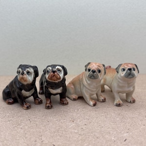 May include: Four ceramic pug dog figurines. Two are black and white, and two are tan and white. The dogs are in various poses, some sitting and some standing. The figurines are detailed and have a glossy finish.