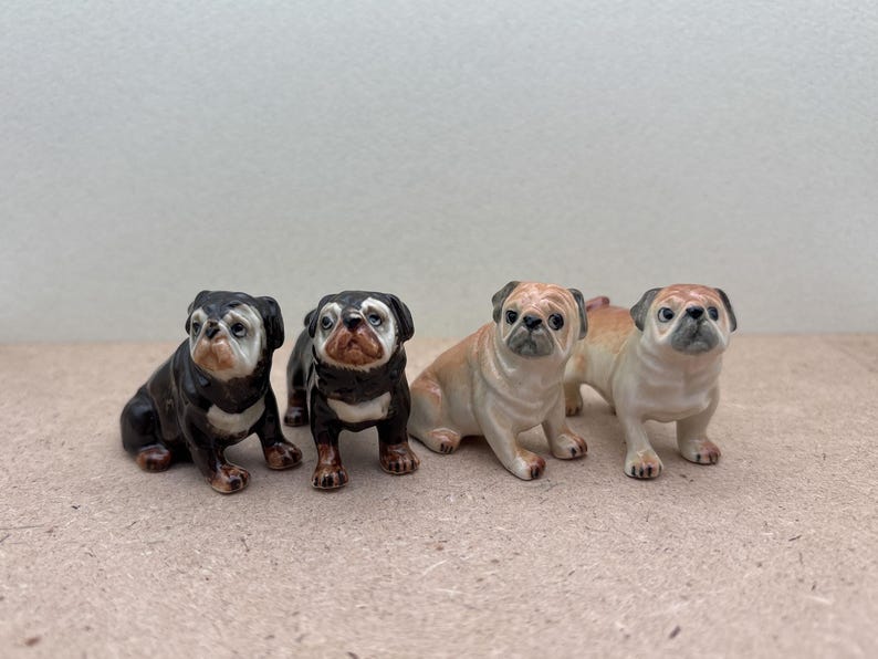 May include: Four ceramic pug dog figurines. Two are black and white, and two are tan and white. The dogs are in various poses, some sitting and some standing. The figurines are detailed and have a glossy finish.