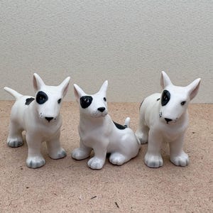 Op de afbeelding: Drie witte bullterriër-beeldjes van keramiek met zwarte markeringen op hun gezicht. De beeldjes staan op een lichtbruine ondergrond.