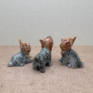 Puede incluir: Tres figuritas de cer&aacute;mica de Yorkshire Terrier. Las figuritas son grises y marrones con detalles negros. Todas miran en la misma direcci&oacute;n, pero cada una tiene una pose ligeramente diferente.