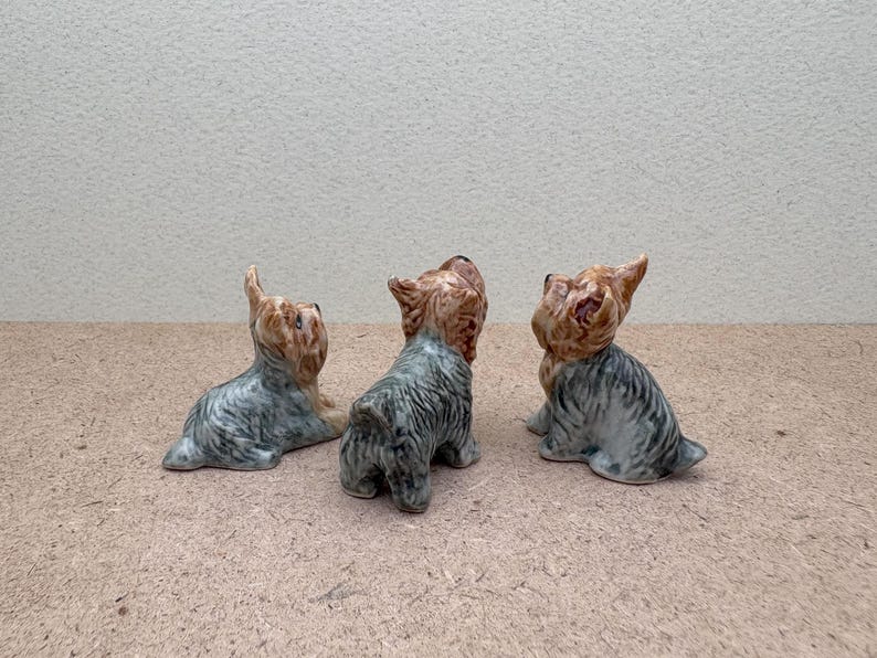 Puede incluir: Tres figuritas de cer&aacute;mica de Yorkshire Terrier. Las figuritas son grises y marrones con detalles negros. Todas miran en la misma direcci&oacute;n, pero cada una tiene una pose ligeramente diferente.