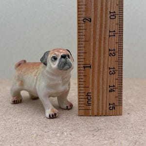 May include: A ceramic pug figurine, tan and cream-colored with a black muzzle and ears, stands next to a wooden ruler. The ruler shows measurements in inches, with the pug approximately 2 inches tall. The figurine has a detailed, realistic appearance.