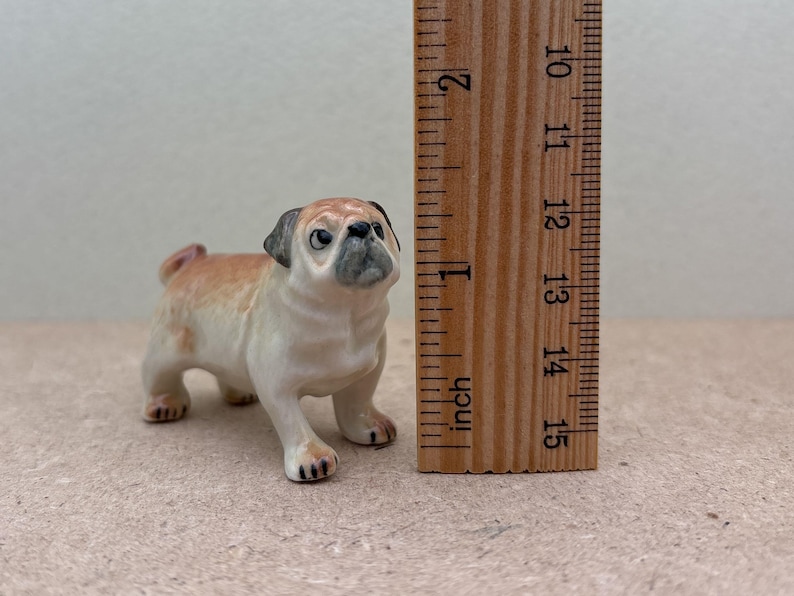 May include: A ceramic pug figurine, tan and cream-colored with a black muzzle and ears, stands next to a wooden ruler. The ruler shows measurements in inches, with the pug approximately 2 inches tall. The figurine has a detailed, realistic appearance.