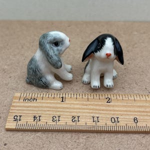 May include: Two small ceramic rabbit figurines, one gray and white with black ears, the other black and white with white ears. The figurines are about 1 inch tall and are sitting on a wooden ruler.