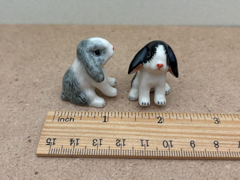 May include: Two small ceramic rabbit figurines, one gray and white with black ears, the other black and white with white ears. The figurines are about 1 inch tall and are sitting on a wooden ruler.