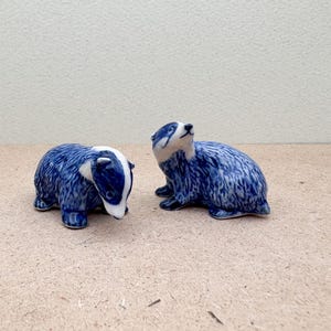 以下が含まれることがあります： 青と白の陶器製のアナグマの置物2つ。1つのアナグマは上を見上げていて、もう1つのアナグマは下を見ている。