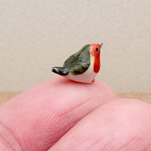 May include: A small, hand-painted porcelain figurine of a robin. The bird is perched on a finger and has a green and gray body with a red breast.