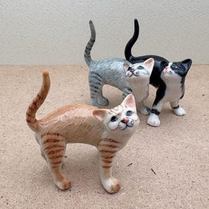 K&ouml;nnte beinhalten: Drei Keramik-Katzenfiguren in verschiedenen Farben. Die vordere Katze ist orange mit braunen Streifen, die anderen sind grau mit Streifen und schwarz-wei&szlig;. Alle Katzen schauen nach oben.
