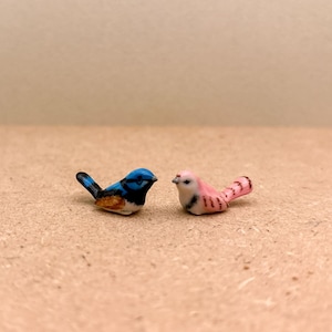 May include: Two small ceramic bird figurines. The bird on the left is blue, black, and white. The bird on the right is pink and white.