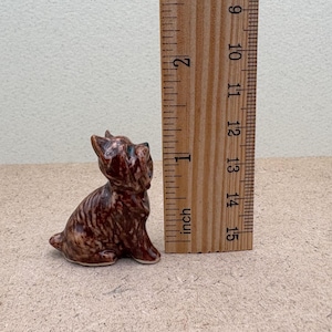 Puede incluir: Una peque&ntilde;a figurita de cer&aacute;mica marr&oacute;n y negra de un perro sentado. La figurita mide aproximadamente 3,8 cm de alto.