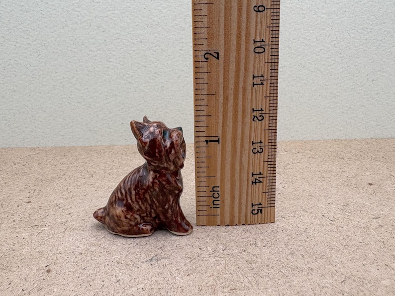 Puede incluir: Una peque&ntilde;a figurita de cer&aacute;mica marr&oacute;n y negra de un perro sentado. La figurita mide aproximadamente 3,8 cm de alto.