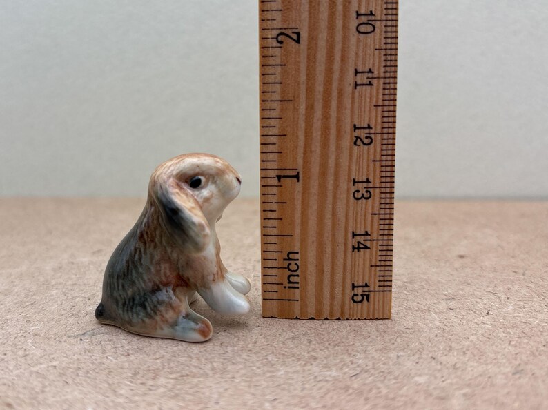 May include: A small ceramic rabbit figurine, about 1.5 inches tall, with brown, black, and white fur. The figurine is sitting on a brown surface next to a ruler.