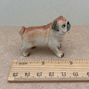 May include: A small ceramic pug figurine, tan and white with black accents. The pug is standing, with its head turned slightly to the side. A wooden ruler is in the foreground, showing measurements in inches.