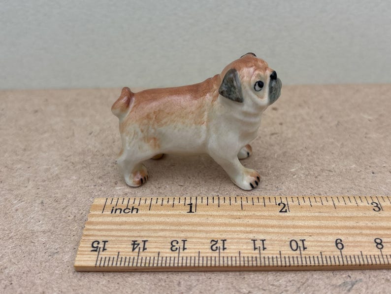May include: A small ceramic pug figurine, tan and white with black accents. The pug is standing, with its head turned slightly to the side. A wooden ruler is in the foreground, showing measurements in inches.