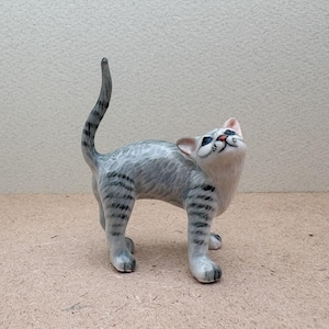 K&ouml;nnte beinhalten: Eine Keramikfigur einer Katze in stehender Pose, die nach oben blickt. Die Katze hat grau-wei&szlig;es Fell mit schwarzen Streifen an Beinen und Schwanz. Die Augen der Katze sind blau, Nase und Mund rosa. Die Figur steht auf einer hellbraunen Oberfl&auml;che.