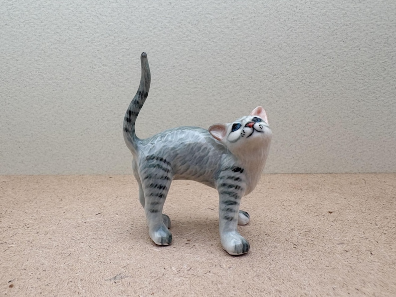 K&ouml;nnte beinhalten: Eine Keramikfigur einer Katze in stehender Pose, die nach oben blickt. Die Katze hat grau-wei&szlig;es Fell mit schwarzen Streifen an Beinen und Schwanz. Die Augen der Katze sind blau, Nase und Mund rosa. Die Figur steht auf einer hellbraunen Oberfl&auml;che.
