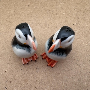 May include: Two ceramic salt and pepper shakers shaped like puffins. The puffins are black and white with orange beaks and feet. They are facing each other.