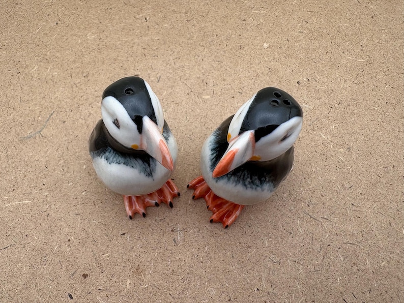 May include: Two ceramic salt and pepper shakers shaped like puffins. The puffins are black and white with orange beaks and feet. They are facing each other.
