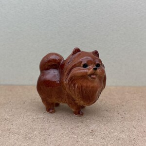 May include: A small ceramic figurine of a brown Pomeranian dog. The dog is standing on all four paws and has a fluffy tail.