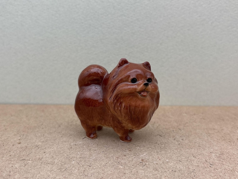 May include: A small ceramic figurine of a brown Pomeranian dog. The dog is standing on all four paws and has a fluffy tail.