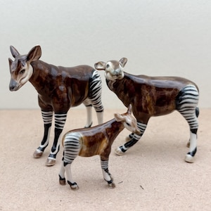 May include: Three ceramic figurines of okapi, a type of African mammal with zebra-like stripes. The figurines are brown and white with black stripes. The figurines are standing on a brown surface.