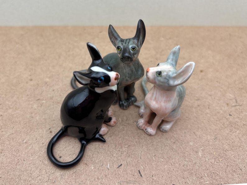 Puede incluir: Tres figuritas de cer&aacute;mica de gatos. Una es negra con marcas blancas, otra es gris con ojos azules y la &uacute;ltima es rosa claro con marcas grises.