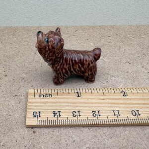 Puede incluir: Una peque&ntilde;a figurita de cer&aacute;mica marr&oacute;n y negra de un perro, de aproximadamente 2,5 cm de alto, de pie sobre una regla de madera.