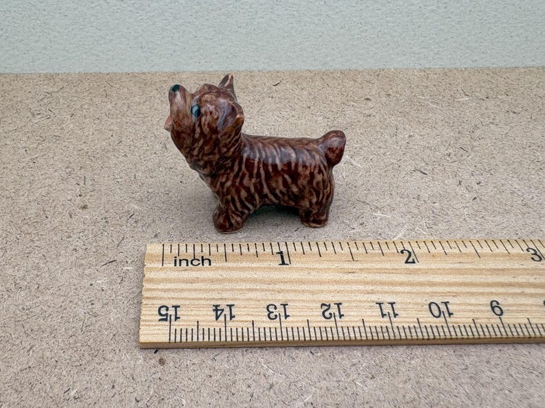 Puede incluir: Una peque&ntilde;a figurita de cer&aacute;mica marr&oacute;n y negra de un perro, de aproximadamente 2,5 cm de alto, de pie sobre una regla de madera.