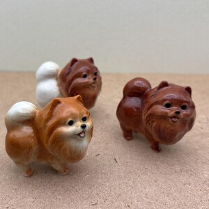 May include: Three ceramic figurines of Pomeranian dogs. The dogs are brown and white and have fluffy tails. The figurines are standing on a brown surface.
