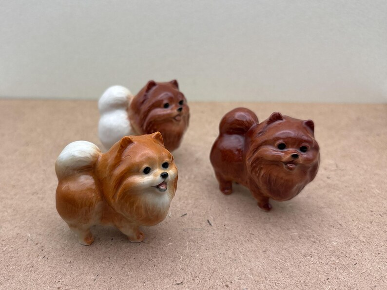 May include: Three ceramic figurines of Pomeranian dogs. The dogs are brown and white and have fluffy tails. The figurines are standing on a brown surface.