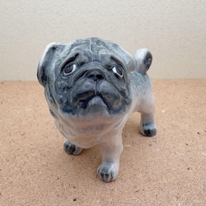 May include: A ceramic pug figurine in shades of gray and white. The dog is depicted in a standing position, with a slightly sad expression. The figurine has detailed features and a glossy finish, suitable for home decor.