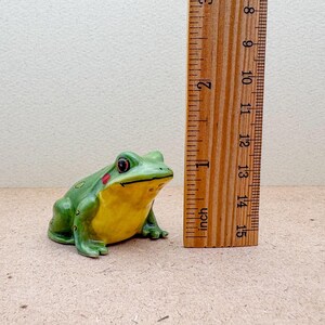 Puede incluir: Una figura de rana de cer&aacute;mica, pintada de verde con vientre amarillo y manchas rojas en las mejillas. La rana mide aproximadamente 10 cm de largo, medido por una regla de madera en la imagen. El objeto es decorativo.