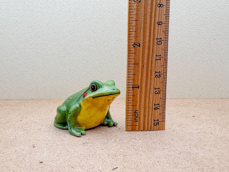 Puede incluir: Una figura de rana de cer&aacute;mica, pintada de verde con vientre amarillo y manchas rojas en las mejillas. La rana mide aproximadamente 10 cm de largo, medido por una regla de madera en la imagen. El objeto es decorativo.