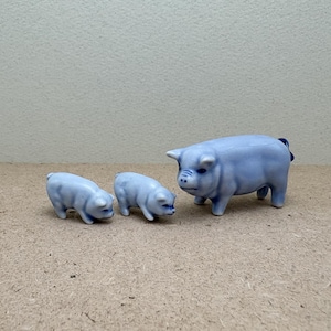 May include: Three light blue ceramic pig figurines. The largest pig stands upright, while two smaller pigs are positioned in front. The figurines have a glossy finish and are set against a neutral background.