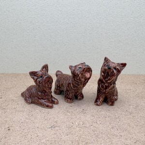 Puede incluir: Tres figuritas de perro de cer&aacute;mica marr&oacute;n. Los perros est&aacute;n en diferentes poses, uno de pie y mirando hacia arriba, otro sentado y mirando hacia adelante, y otro acostado y mirando hacia un lado.