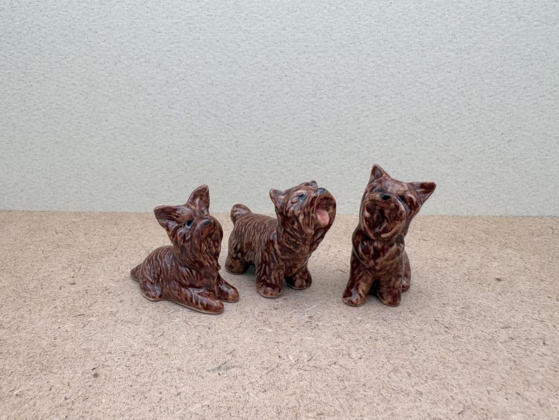 Puede incluir: Tres figuritas de perro de cer&aacute;mica marr&oacute;n. Los perros est&aacute;n en diferentes poses, uno de pie y mirando hacia arriba, otro sentado y mirando hacia adelante, y otro acostado y mirando hacia un lado.