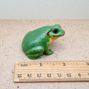 Puede incluir: Figura de rana de cer&aacute;mica verde, con detalles amarillos y rojos, colocada sobre una superficie marr&oacute;n claro. La rana mide unos 5 cm de largo. Una regla de madera est&aacute; al lado.