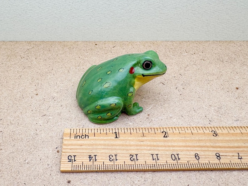 Puede incluir: Figura de rana de cer&aacute;mica verde, con detalles amarillos y rojos, colocada sobre una superficie marr&oacute;n claro. La rana mide unos 5 cm de largo. Una regla de madera est&aacute; al lado.