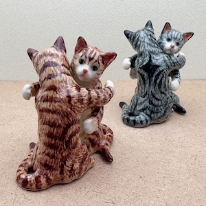 May include: Two ceramic cat figurines in a hugging pose. One is brown with dark brown stripes, the other is gray with black stripes. Both have white paws and are hugging a smaller cat with a pink nose.