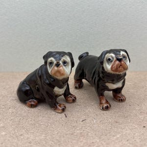 May include: Two ceramic bulldog figurines. One is sitting, the other is standing. Both have black and white fur with brown accents on their paws and faces. The figurines are detailed and appear to be hand-painted.