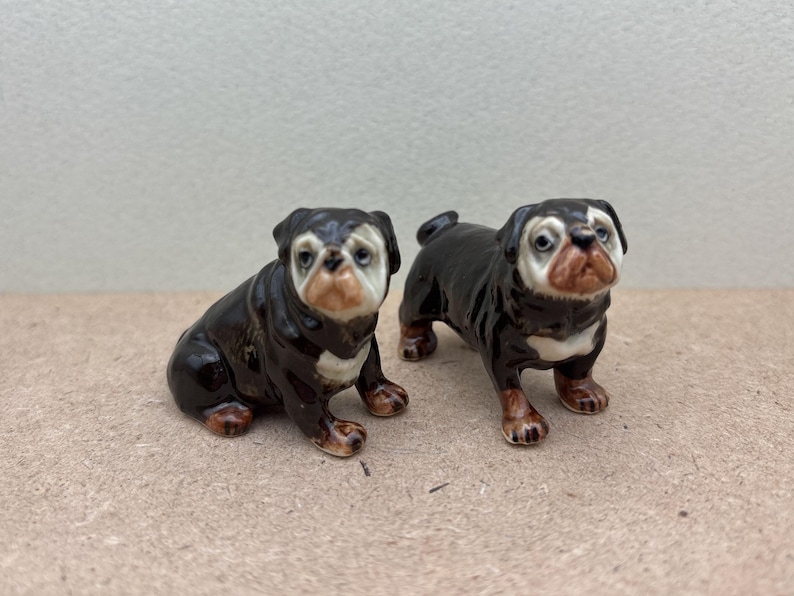 May include: Two ceramic bulldog figurines. One is sitting, the other is standing. Both have black and white fur with brown accents on their paws and faces. The figurines are detailed and appear to be hand-painted.