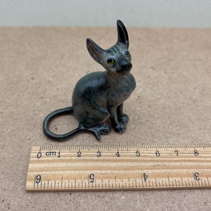 Puede incluir: Una peque&ntilde;a figurita de cer&aacute;mica de un gato gris y negro sentado con la cola enroscada alrededor de su cuerpo. La figurita mide aproximadamente 10 cm de alto.