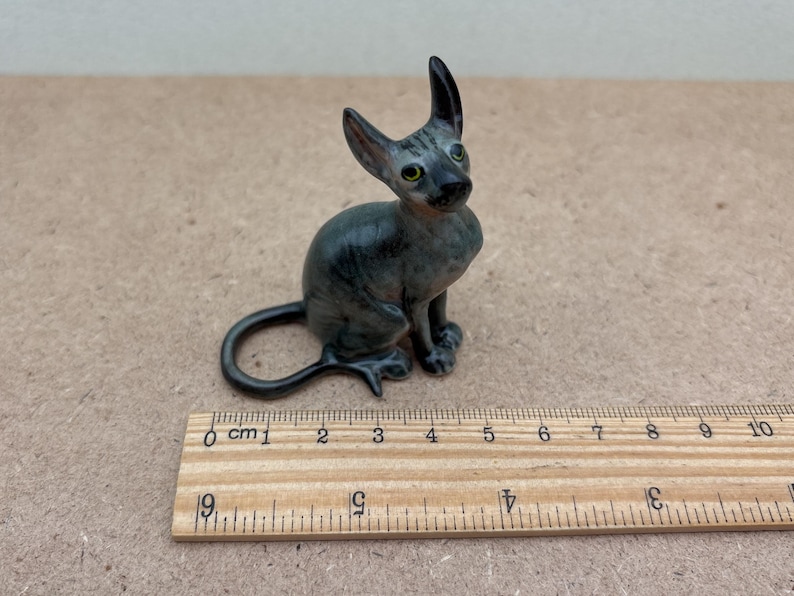 Puede incluir: Una peque&ntilde;a figurita de cer&aacute;mica de un gato gris y negro sentado con la cola enroscada alrededor de su cuerpo. La figurita mide aproximadamente 10 cm de alto.