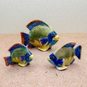 May include: Three ceramic fish figurines in blue, green and yellow colours. The fish are of different sizes and have detailed painted scales and fins.