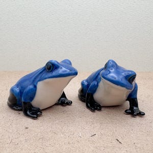 May include: Two ceramic frog ornaments. The frogs are blue with white bellies and black feet. They are posed as if they are sitting. The background is a light beige colour.
