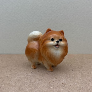 May include: A small, ceramic figurine of a brown and white Pomeranian dog. The dog is standing on all four paws and has a fluffy tail.